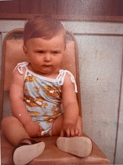 A serious toddler sits on a chair outside.