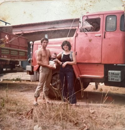 Baby Fabiano is held by his father in front of a tractor with his mother next to them.